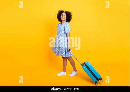 Tutta la lunghezza di una bella ragazza afro-americana o brasiliana felice con capelli ricci, in un abito estivo blu, un viaggiatore, si erge su uno sfondo giallo isolato con una valigia blu, guarda via, sorride Foto Stock