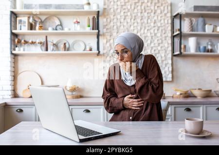 Una giovane araba in hijab è incinta e lavora su un computer portatile. Si sente male, sente forte nausea, sollecita a vomitare, copre la bocca con la mano, sintomi di gravidanza. Foto Stock