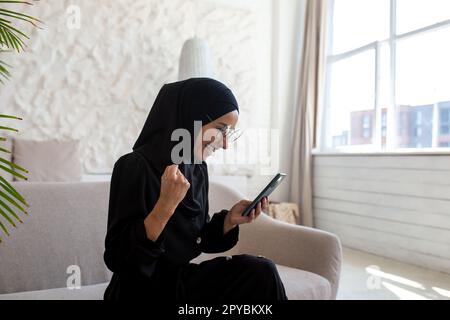 Giovane donna musulmana in hijab nero e burqa seduto sul divano a casa e utilizzando il telefono. Addicted ai giochi in linea, ai posti scommettono, alle buone notizie ricevute, ai messaggi. Mostra un gesto di vittoria con la mano. Foto Stock