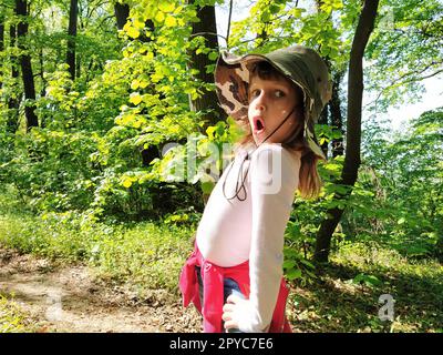 la bella ragazza di 6 anni con un cappello da caccia si fa una grappa sul viso. Emozione di sorpresa, paura o scoraggiamento. Il bambino si gira e guarda la telecamera. Foresta sullo sfondo Foto Stock