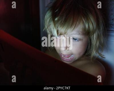 Una bambina carina gioca con un tablet. Un bambino di 7 anni con un viso sorridente e capelli biondi guarda in un gadget. Copia spazio. Ragazza nella stanza. Parete scura e chiara. A casa la sera Foto Stock