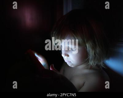 Una bambina carina gioca con un tablet. Un bambino di 7 anni con una faccia seria e capelli biondi guarda in un gadget. Copia spazio. Ragazza nella stanza. Parete scura e chiara. A casa di notte Foto Stock