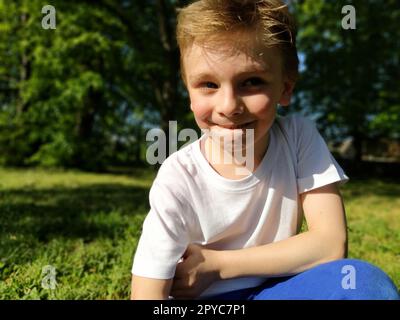 Un giovane ragazzo che sorride e guarda di lato. Il bambino indossa una T-shirt bianca e pantaloni blu. I capelli biondi sono bellissimi cadono sulla fronte. Ciglia bionda lunghe. Parcheggiare sullo sfondo. Foto Stock