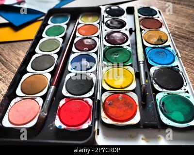 Una serie di acquerelli di colori diversi per il disegno. Contenitori rotondi multicolori con vernice lucida. Strumenti per la lezione di disegno e manodopera. Carta colorata. Rosso, blu, giallo, verde. Foto Stock