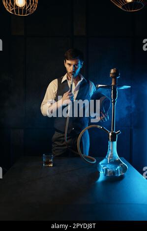 Ragazzo vestito retro che fuma hookah mentre si trova in piedi tra nuvole di fumo Foto Stock