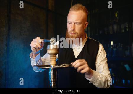 Uomo elegante barbuto che mescola tabacco in narghilè e fumo Foto Stock