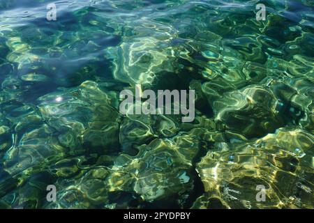 Consistenza di acqua salata trasparente blu turchese verde. Vista dall'alto della superficie dell'acqua e delle increspature. Acqua onde di mare sullo sfondo. Il fondo roccioso con alghe è visibile attraverso l'acqua cristallina Foto Stock