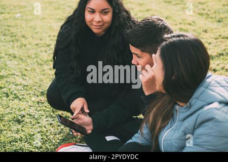 La famiglia ispanica trascorre del tempo di qualità insieme in un parco locale in una splendida giornata di sole. Un ragazzo adolescente è seduto sull'erba e tiene lo smartphone mentre guarda lontano. Sua madre e sua sorella sono sedute accanto a lui, guardando la telecamera e sorridendo. Foto Stock