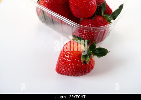 Fragole mature in una confezione di plastica su sfondo bianco. Deliziose bacche fresche in un contenitore per la vendita ai clienti. Mantenere il cibo fresco. Cibo sano, una fragola estratta dal contenitore Foto Stock