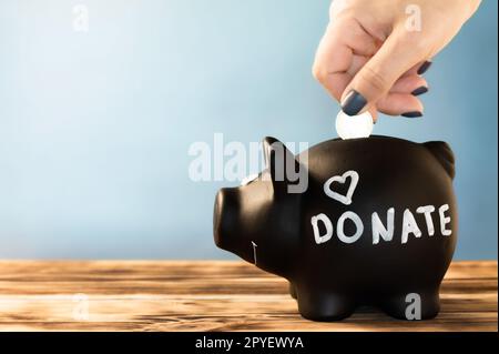 Mano mettendo una moneta in un salvadanaio nero con un cartello donate a gesso su sfondo blu. Concetto di donazione e beneficenza Foto Stock