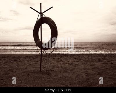 Lifebuoy su una spiaggia sabbiosa. Cerchio arancione su un palo per salvare le persone che annegano nel mare. Punto di salvataggio sulla riva. Cielo e mare sullo sfondo. Seppia monocromatica Foto Stock