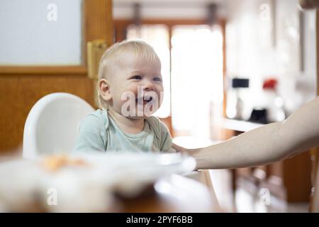 Adorabile allegro e felice bambino che sorride seduto in seggiolone al tavolo da pranzo in cucina a casa, bevendo un cucchiaio alimentato da sua madre Foto Stock