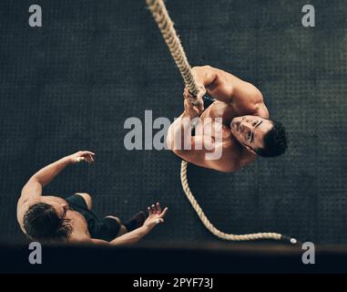 Quasi là. Colpo ad angolo alto di una forma e determinato giovane uomo che si arrampica su una corda per l'esercizio di forza in una palestra. Foto Stock