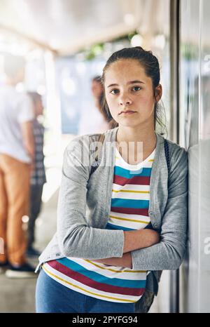 La scuola può essere un luogo solitario. Ritratto di una studentessa solitaria che si trova fuori dalla sua classe con compagni di classe sullo sfondo. Foto Stock