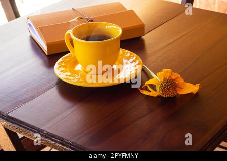 Goiânia, Goias, Brasile – 01 maggio 2023: Una tazza gialla piena di caffè, con un piattino e un quaderno, su un tavolo di legno, con un girasole. Foto Stock