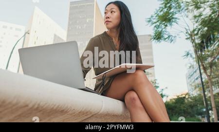 La ragazza giovane usa il PC portatile fa note nel notebook dall'esterno sullo sfondo degli edifici moderni Foto Stock