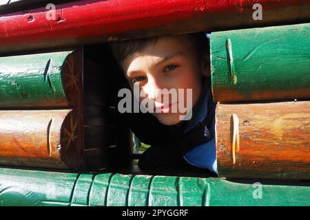 Un ragazzo caucasico di 10 anni con capelli biondi e occhi grigi guarda fuori dalla finestra di una casa di legno su un parco giochi per bambini. Peekaboo. Il bambino si è nascosto. Bella faccia Foto Stock