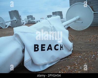 Un ombrellone è stato ripiegato su una spiaggia sabbiosa dopo il maltempo. Tempesta in mare. Fine della stagione balneare a causa del tifone e del ciclone. Lettini, ombrelloni e mobili dell'hotel. Parola sulla spiaggia scritta su bianco Foto Stock