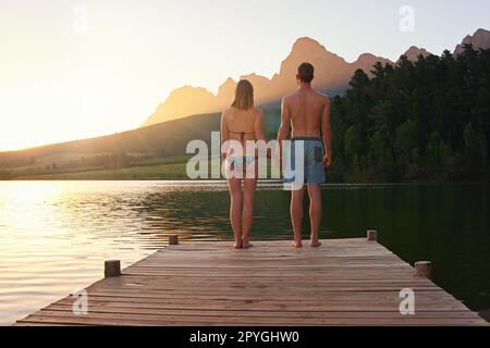 Un tramonto perfetto. Ripresa da dietro di una giovane coppia in costume da bagno in piedi su un molo al tramonto. Foto Stock