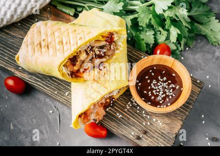 Arrotolare lo shawarma con semi di sesamo, salsa barbecue, pollo in pita su una superficie di legno con pomodori ciliegini e prezzemolo Foto Stock