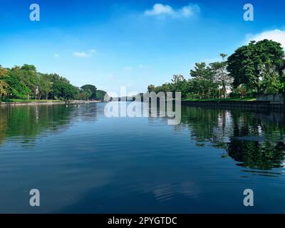 Una grande piscina nel mezzo del parco è piena di grandi alberi circondati dal cielo blu luminoso Foto Stock