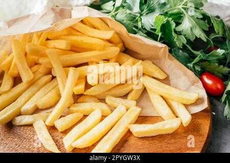 Ampia porzione di patatine fritte in carta artigianale con pomodori ciliegini e prezzemolo verde, su superficie di legno Foto Stock