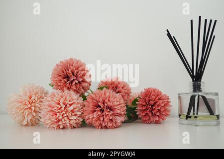 Composizione con bastoncini di incenso, olio essenziale in vaso di vetro e fiori artificiali di spugna rosa all'interno di una stanza bianca. Sfondo bianco. Molti fiori e bastoni profumati. Foto Stock