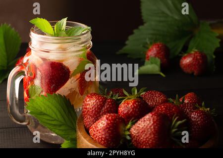 Bevanda fredda estiva con menta, ghiaccio, limone e fragole e ciotola in legno di fragole fresche su superficie di legno nero. Foto Stock