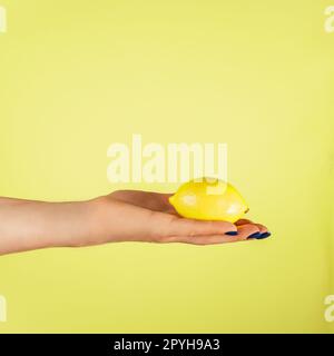 Mano femminile con unghie blu che reggono il limone fresco isolato sul giallo. Spremere il succo. Argomento di dieta. Verticale. Copia spazio Foto Stock