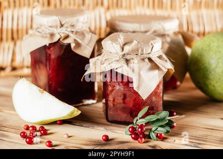 Barattoli di lingonberry e marmellata di pera fatti in casa con carta artigianale sui coperchi su un tavolo di legno accanto a lingonberry e pere fresche Foto Stock