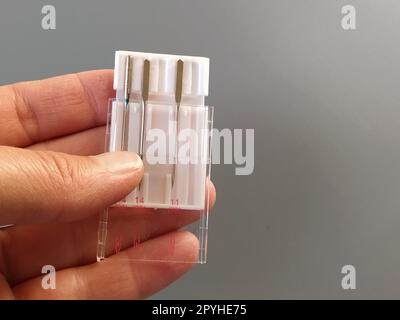 un set di aghi per macchine da cucire in una custodia di plastica bianca. La mano di una donna tiene gli aghi per una macchina da cucire elettrica. Sfondo grigio. Spessori dell'ago diversi Foto Stock