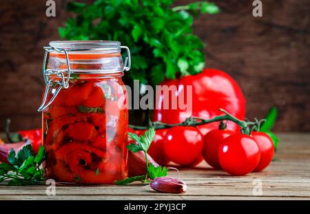 Peperone rosso paprika al forno in vaso di vetro, marinato con peperoncino, aglio ed erbe aromatiche. Pomodori freschi, prezzemolo, baguette. Rustico tavolo da cucina in legno backgrou Foto Stock