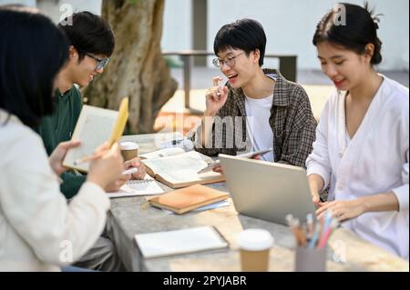 Giovani studenti asiatici maschi giovani felici e intelligenti che aiutano e tutorano l'economia ai suoi amici in un campus Park. Foto Stock