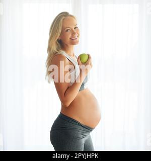 Alcuni lo chiamano mangiare sano, io lo chiamo amore. Ritratto di una donna felice incinta vestita di abbigliamento sportivo e mangiare una mela a casa. Foto Stock