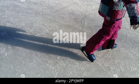 I bambini viaggiano in un parco cittadino su una pista di pattinaggio su ghiaccio. Gambe e mani di uno skater mentre pattina sul ghiaccio. La ragazza è vestita con abbigliamento sportivo rosa. Pattini blu vestiti sulle gambe. Foto Stock