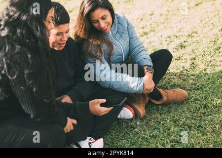 La famiglia ispanica trascorre del tempo di qualità insieme in un parco locale in una splendida giornata di sole. Un ragazzo adolescente è seduto sull'erba e tiene lo smartphone mentre guarda lontano. Sua madre e sua sorella sono sedute accanto a lui, guardando la telecamera e sorridendo. Foto Stock