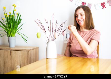 Donna sorridente che decora un bouquet di rami di salice con uova di Pasqua colorate a casa. Buona Pasqua Foto Stock