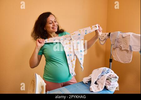 Giovane donna incinta con grande pancia stand da tavolo da stiro a casa, sorrisi, tenendo il bodysuit neonato e smistamento vestiti Foto Stock