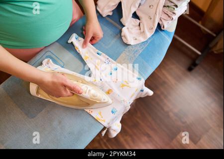 Vista dall'alto di una donna incinta in piedi accanto all'asse da stiro e muta per bambini fumante con ferro a vapore elettrico. Foto Stock