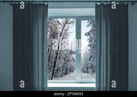 Finestra in una camera con vista sul paesaggio invernale. Camera con finestra. Finestra con tende grigie. Tempo freddo e nevoso fuori dalla finestra. Foto Stock