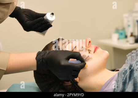 Il cosmetologo applica il nanoogel di carbonio del volto del cliente. Preparazione per il trattamento laser della pelle. Pelatura del viso in carbonio. Cosmetologia hardware Foto Stock