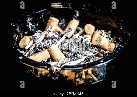 Overhead di bruciare sigaretta in posacenere su sfondo nero. Foto che mostra una terribile dipendenza che causa molte malattie. Foto con profondità ridotta di Foto Stock