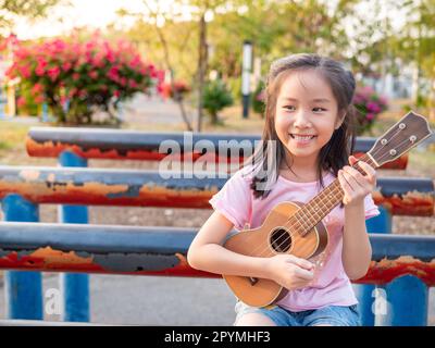 Bambina asiatica giocare l'ukulele, nel giardino sul tubo di acciaio Foto Stock