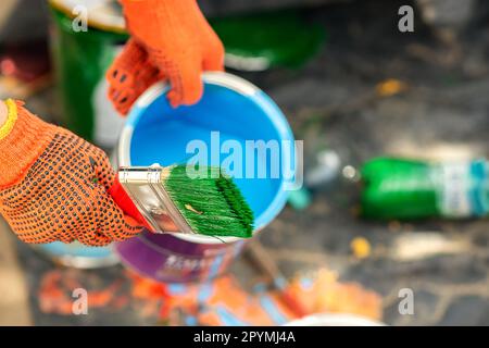 mani nei guanti di protezione arancioni, nelle mani di un secchio con vernice blu e di una spazzola rossa con setole verdi, verniciare la superficie con vernice blu. Foto Stock