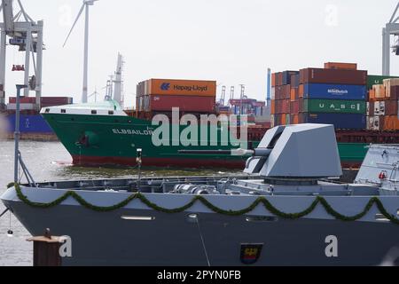 Amburgo, Germania. 04th maggio, 2023. Una nave container naviga durante la cerimonia di denominazione della corvette F266 'Emden' (davanti), seconda di un totale di cinque nuove navi per la Marina, presso il cantiere navale Blohm Voss nel porto. Circa sei anni dopo l'ordine di costruzione, la seconda delle cinque nuove navi navali viene battezzata. La corvette della Marina tedesca è ufficialmente chiamata 'Emden'. Tutte e cinque le nuove cornici sono in costruzione in diversi cantieri navali della Germania settentrionale sotto la direzione del Gruppo NVL. Credit: Marcus Brandt/dpa/Alamy Live News Foto Stock