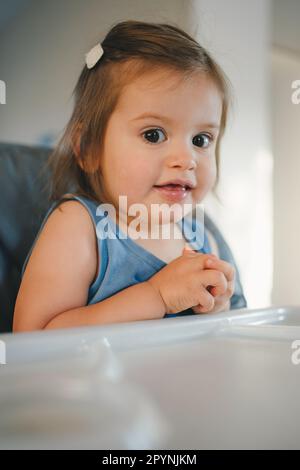 Vista ravvicinata di un bambino seduto in un seggiolone per mangiare in cucina, guardando la c amera e sorridendo. Ritratto di un bambino carino felice Foto Stock