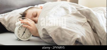 ragazzo sdraiato sul letto e la sveglia di mattina. la mano dei bambini arriva per la sveglia Foto Stock