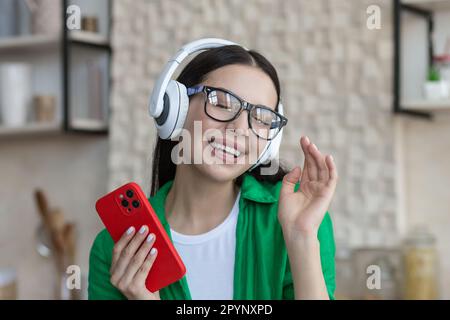 Primo piano. Una giovane studentessa sorridente ascolta la musica in cuffia a casa, tiene un telefono in mano. Chiuse gli occhi, riposa, gode, balla. Foto Stock