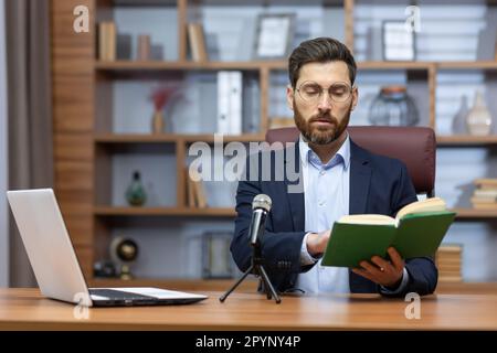 Apprendimento a distanza. Un giovane insegnante maschile siede in ufficio a un tavolo di fronte a un computer portatile e a un microfono e legge una lezione online da un libro a studenti, studenti. Foto Stock