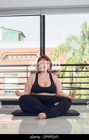 immagine verticale giovane più dimensione donna latina, facendo yoga meditando con gli occhi chiusi in posizione lotus, in interni, indossando abbigliamento sportivo nero. Foto Stock